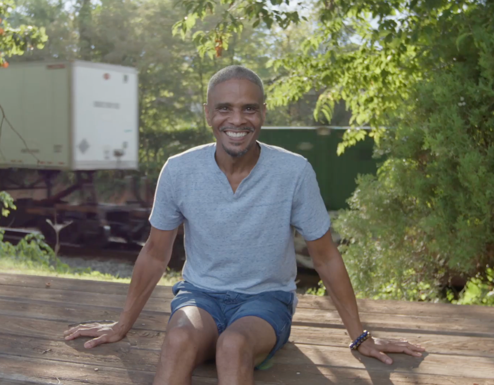 older black man seated on a wood table in front of trees and a semi truck.