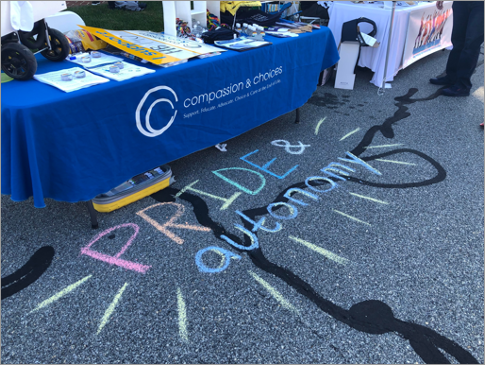 PRIDE & autonomy written in rainbow colored chalk in front of Compassion & Choices booth at a street festival