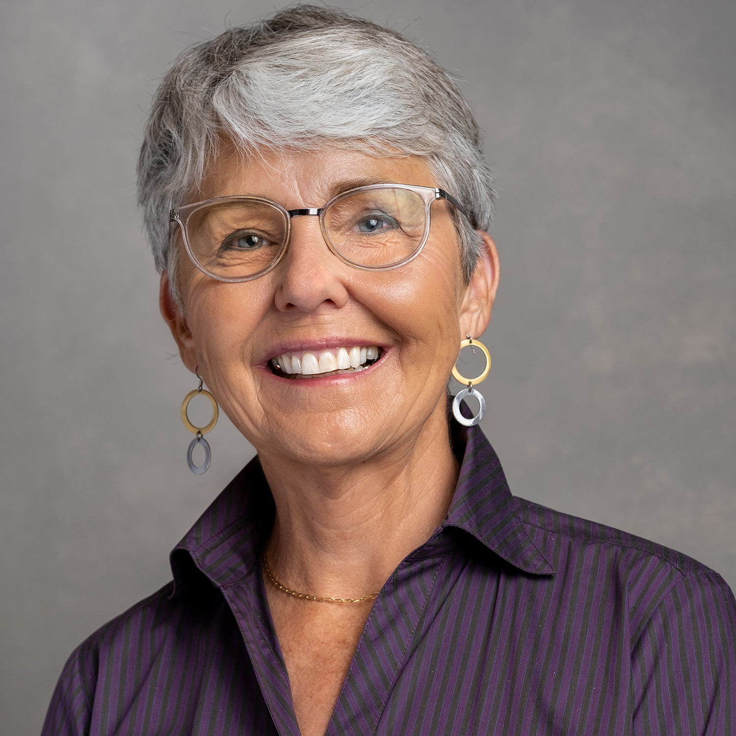 Kim Martin wearing a purple shirt, gold and silver hoop earrings, and smiling in front of a grey backdrop.