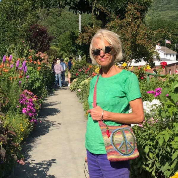 Jill Hammerberg CT Storyteller stands in a garden of colorful flowers wearing a bright green tshirt, sunglasses and a purse with a peace sign.